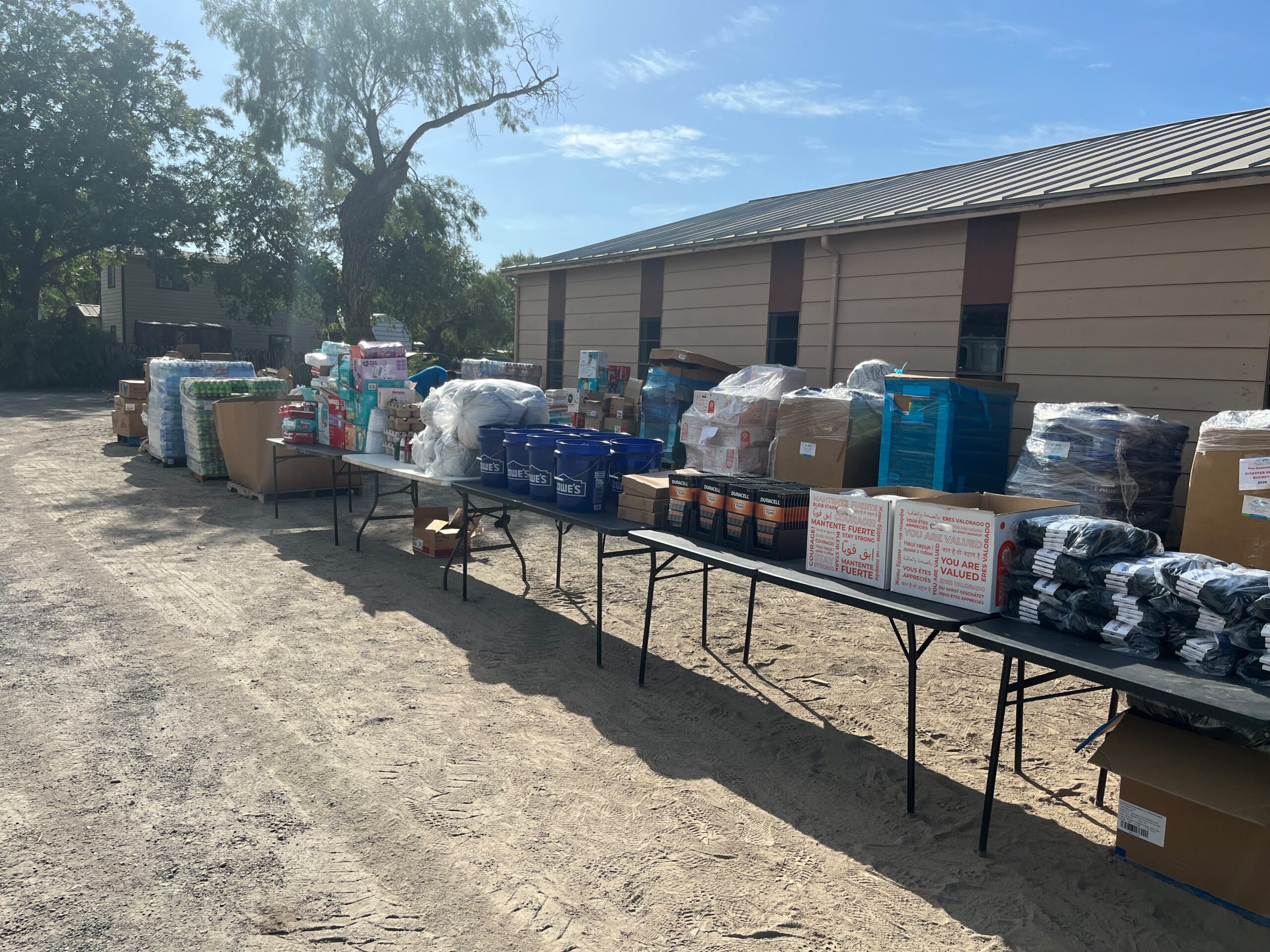 Rows of folding tables loaded with diapers, hygiene buckets, batteries, and blankets staged for distribution in Uvalde