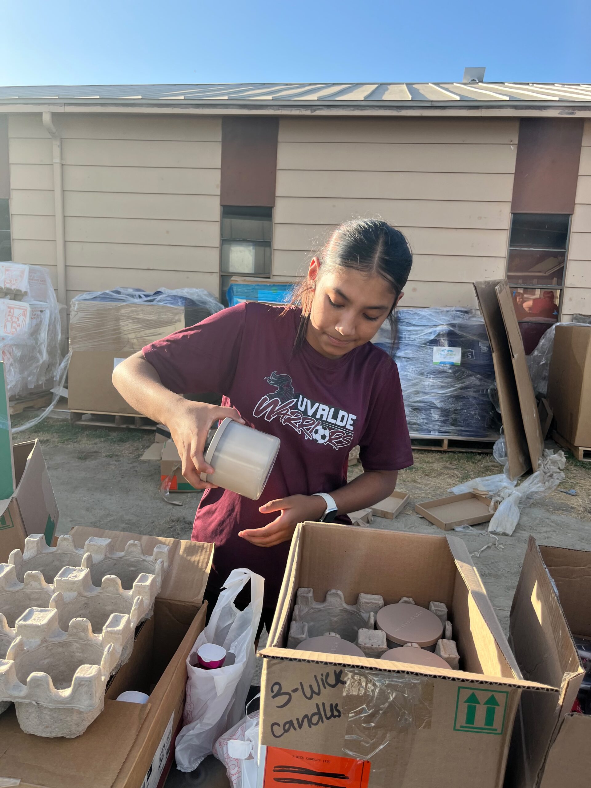 A volunteer in an Uvalde Warriors shirt packing candles into a cardboard box labeled 3-Wick Candles