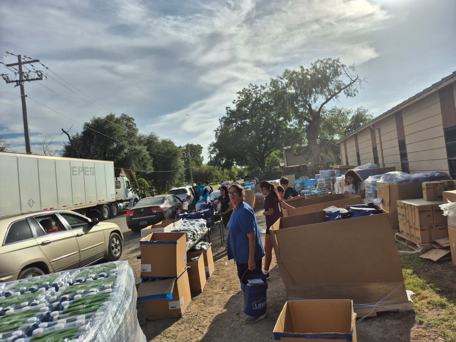 A line of cars waiting along a dirt road next to a trailer marked EPES, with volunteers working a supply table